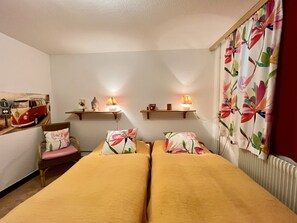 Una stanza con due letti, una sedia, uno scaffale con oggetti decorativi e una finestra con tende floreali.