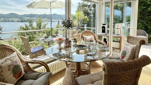 A sunlit terrace with wicker furniture, a glass table, and a view of a lake and hills. - Seepanorama by Interhome (Öhningen)