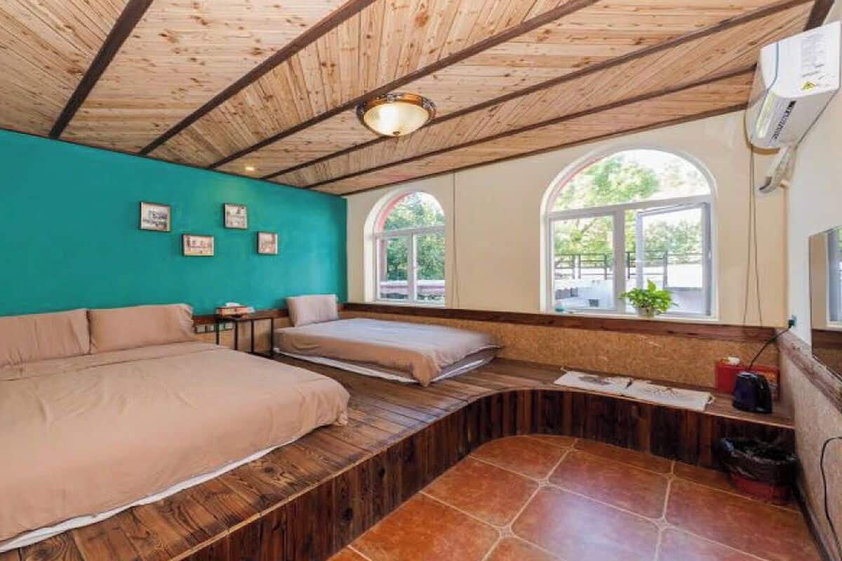 A room with a wooden ceiling, a teal wall, two beds, a bench, and a window with a view.
