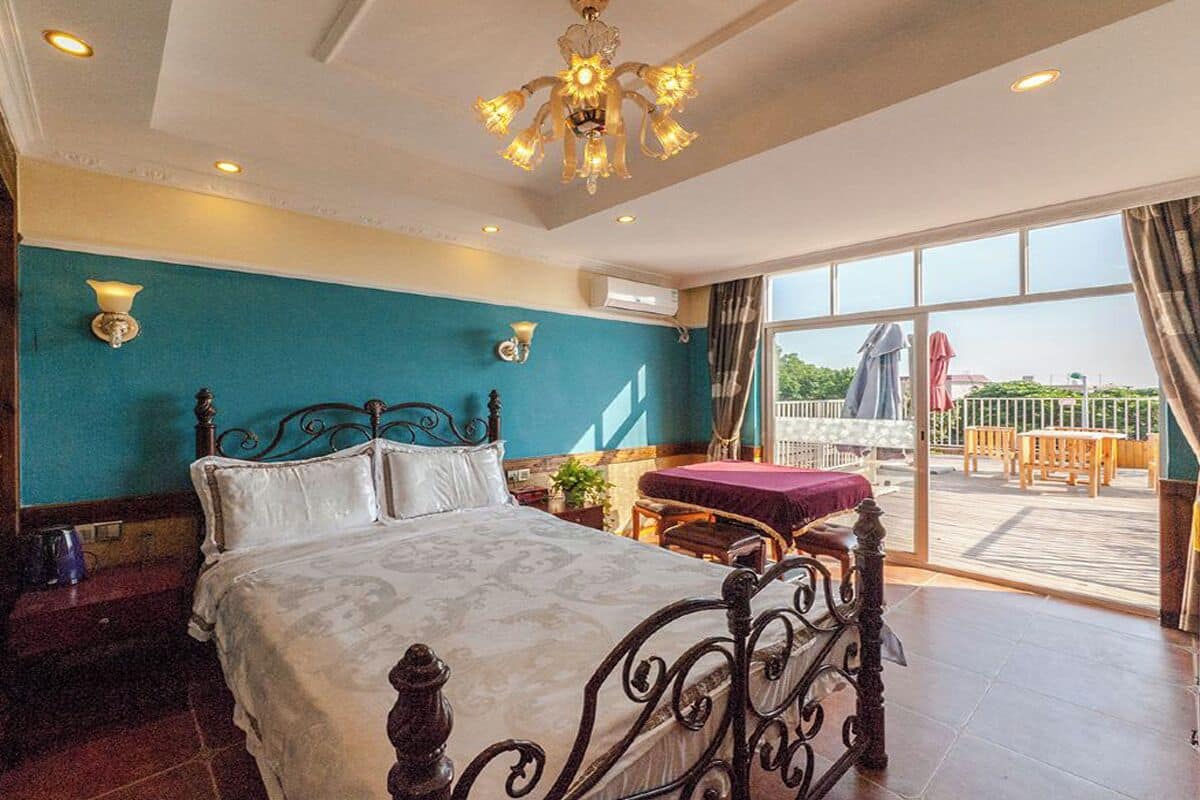 A bedroom with a large bed, a chandelier, a balcony with outdoor furniture, and a view of the outdoors.