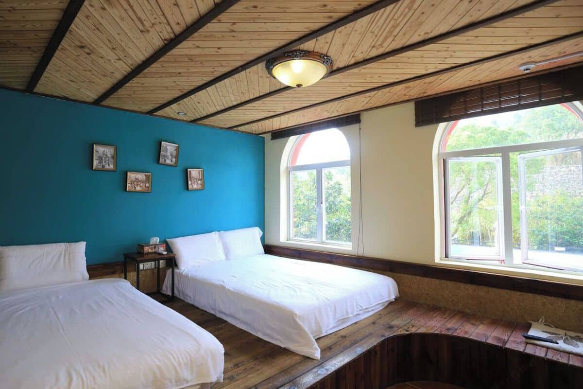 A room with two beds, a wooden ceiling, a large window, and framed pictures on the wall.