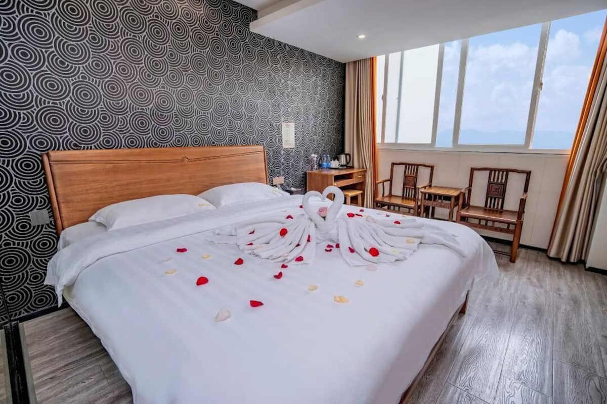 Ein Hotelzimmer mit einem großen Bett, einem hölzernen Kopfteil, einem Schreibtisch mit Stuhl und Blick zum Himmel durch große Fenster.