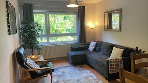 A cozy living room with a dark grey sofa, a wooden chair, a small table with a lamp, and a large window with curtains.