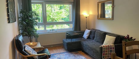 Una sala de estar acogedora con un sofá gris oscuro, una silla de madera, una mesita con lámpara y un ventanal con cortinas.