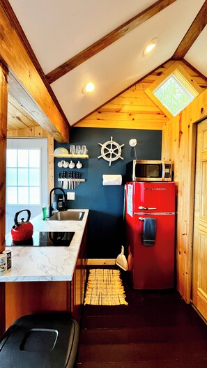 A compact kitchen with a wooden ceiling, a red refrigerator, a microwave, and a ship's wheel decoration.