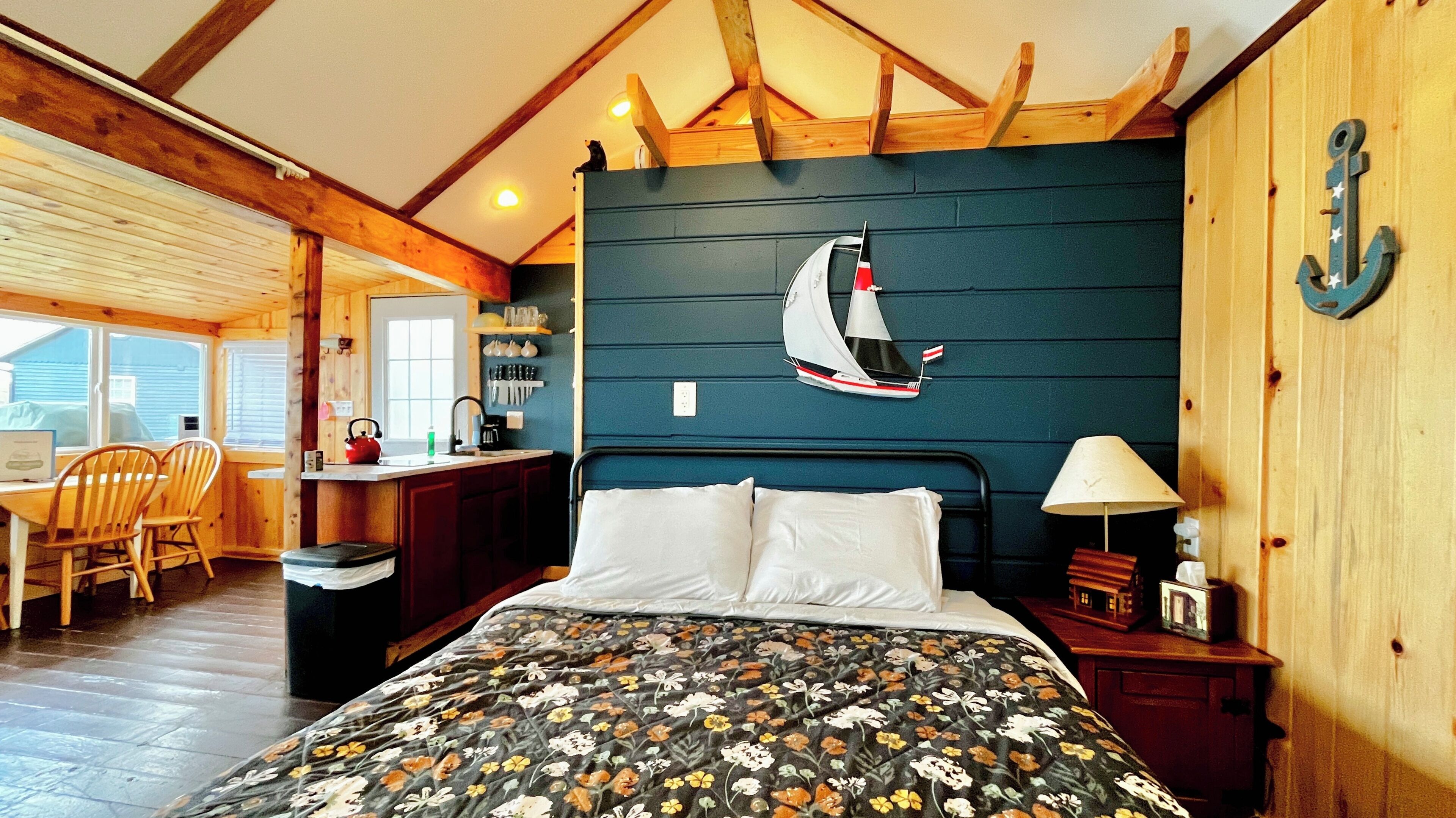 A bedroom with a sailboat wall art, a bed with floral bedding, a desk with chairs, and a view of the outdoors.