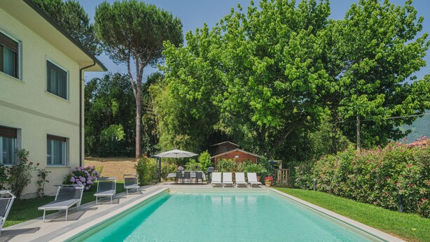 A swimming pool with lounge chairs and a barbecue area, surrounded by lush greenery and a building. - Gerry House by Interhome (Camaiore)