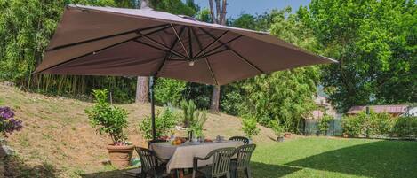 An outdoor patio with a table and chairs under a large umbrella, surrounded by greenery and trees.