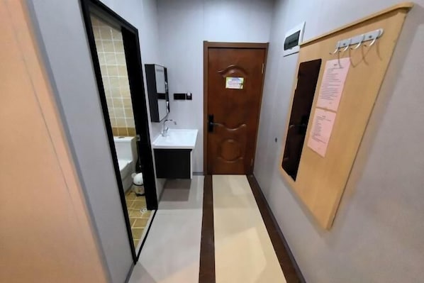 Un couloir étroit, avec une salle de bain sur la gauche, une porte en bois avec un tableau d’affichage sur la droite, et un sol carrelé.