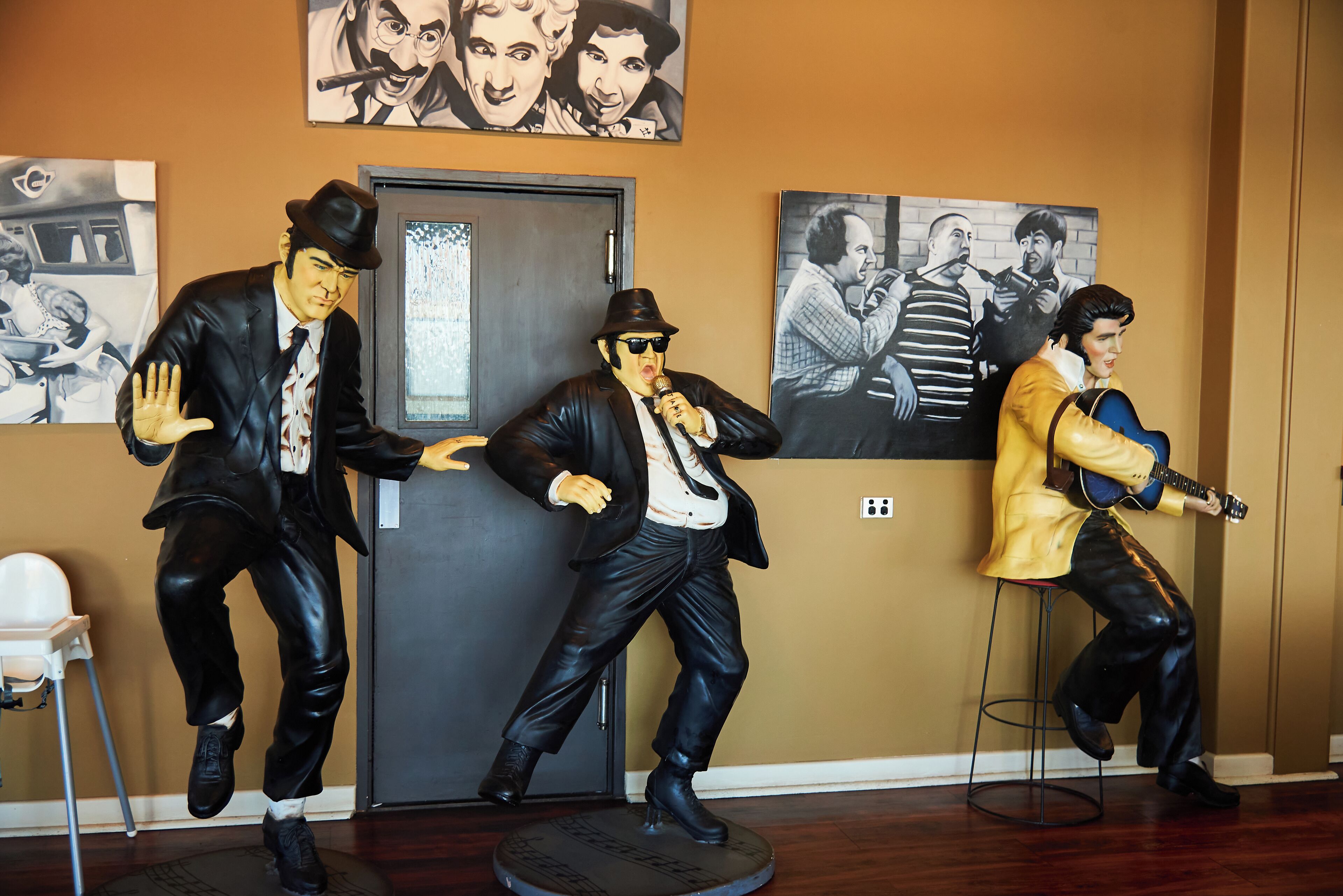 Tres estatuas de hombres con trajes y sombreros, uno tocando una guitarra, ubicadas en una habitación con una puerta y cuadros en la pared.