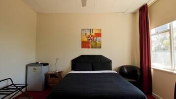 Quarto de hotel com cama, escrivaninha, cadeira, pequeno frigorífico e janela com cortinas.