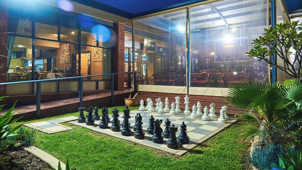 A chessboard set up in a courtyard with a glass enclosure and a brick building in the background. - Mirambeena Motel (Whyalla)
