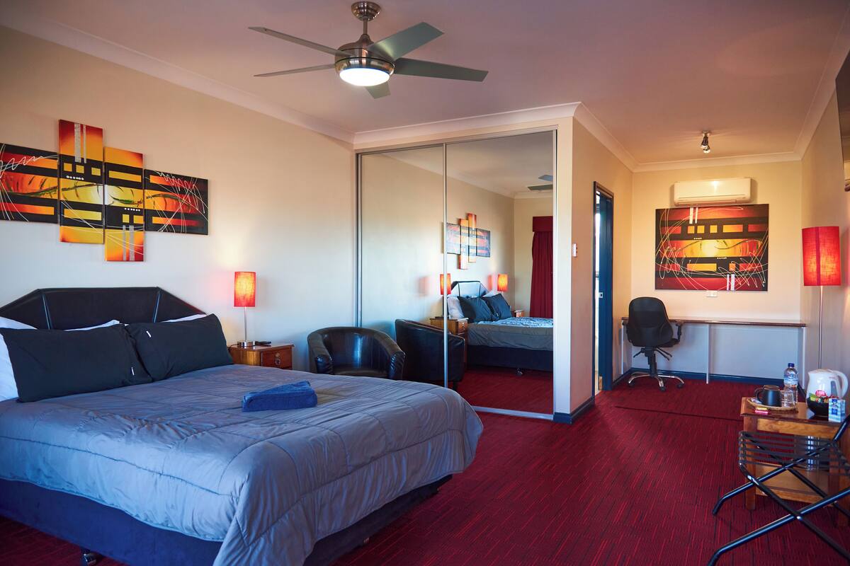 Une chambre d’hôtel avec un grand lit, un bureau, une chaise, un ventilateur de plafond et des œuvres d’art accrochées aux murs.