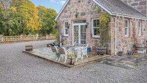 A stone building with a gravel driveway, a patio with outdoor seating, and a view of trees with autumn foliage. - The Doune Luxury Highland House, hot tub, sleeps 12 (Inverness)