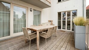 Een houten terras met een tafel en stoelen, grote schuifdeuren en een witte gevel.