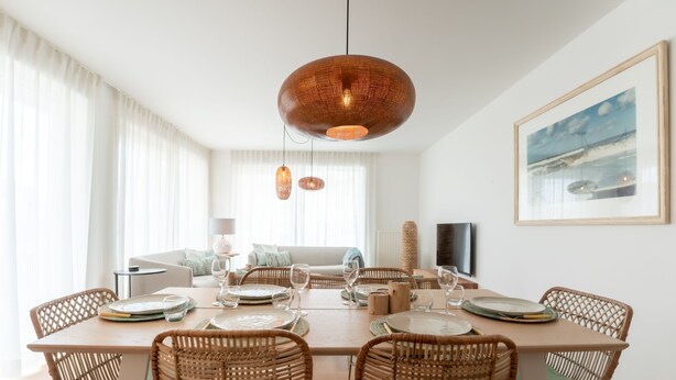 Een eethoek met een grote tafel, rieten stoelen en een hangende koperen lamp.