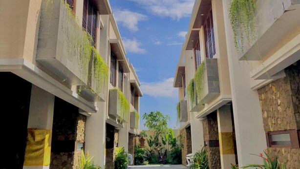 Un pasillo flanqueado por edificios, con balcones adornados con plantas, un muro de piedra a la derecha y un cielo despejado.