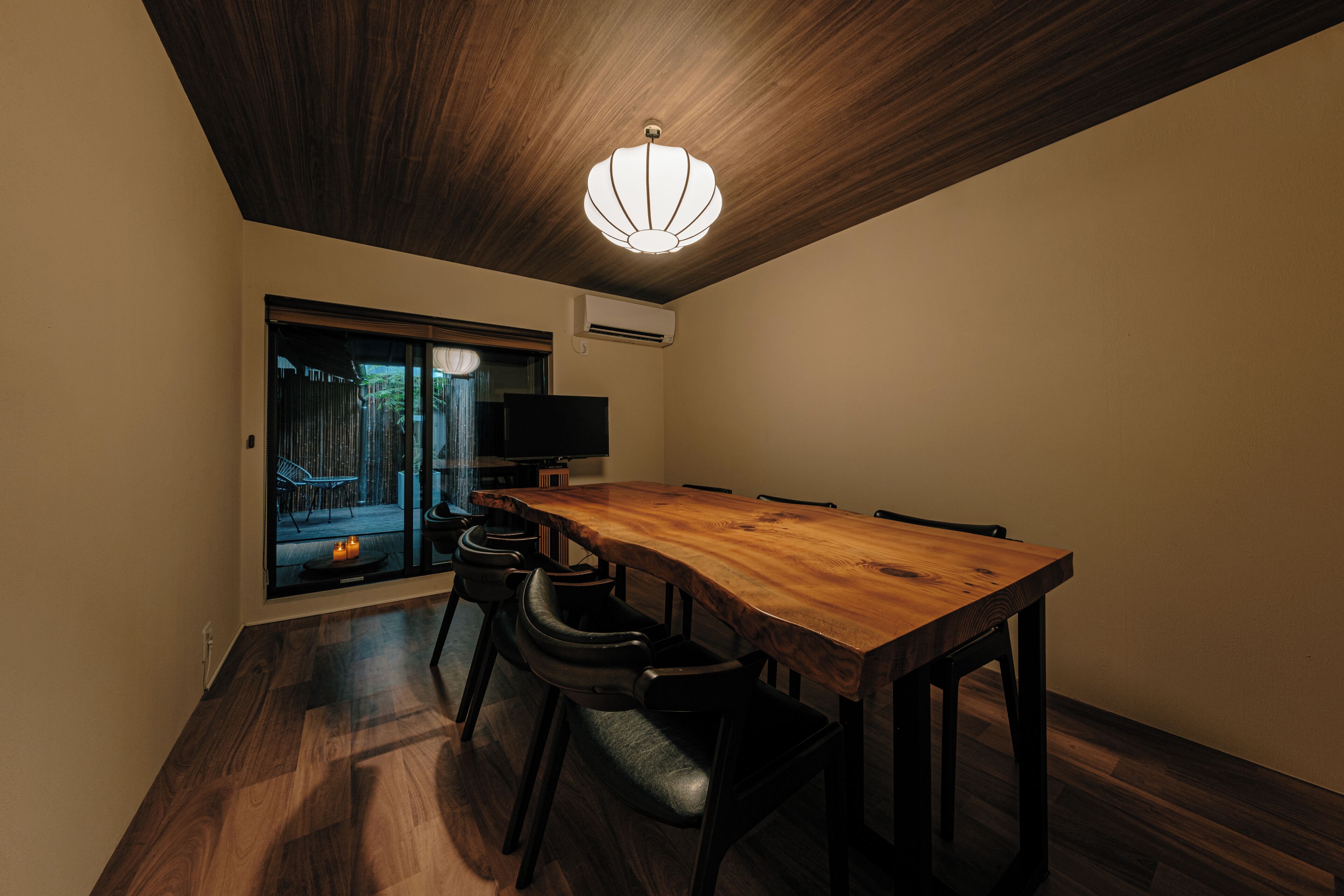 A dining area with a wooden table and chairs, a ceiling light fixture, and a view through a glass door.
