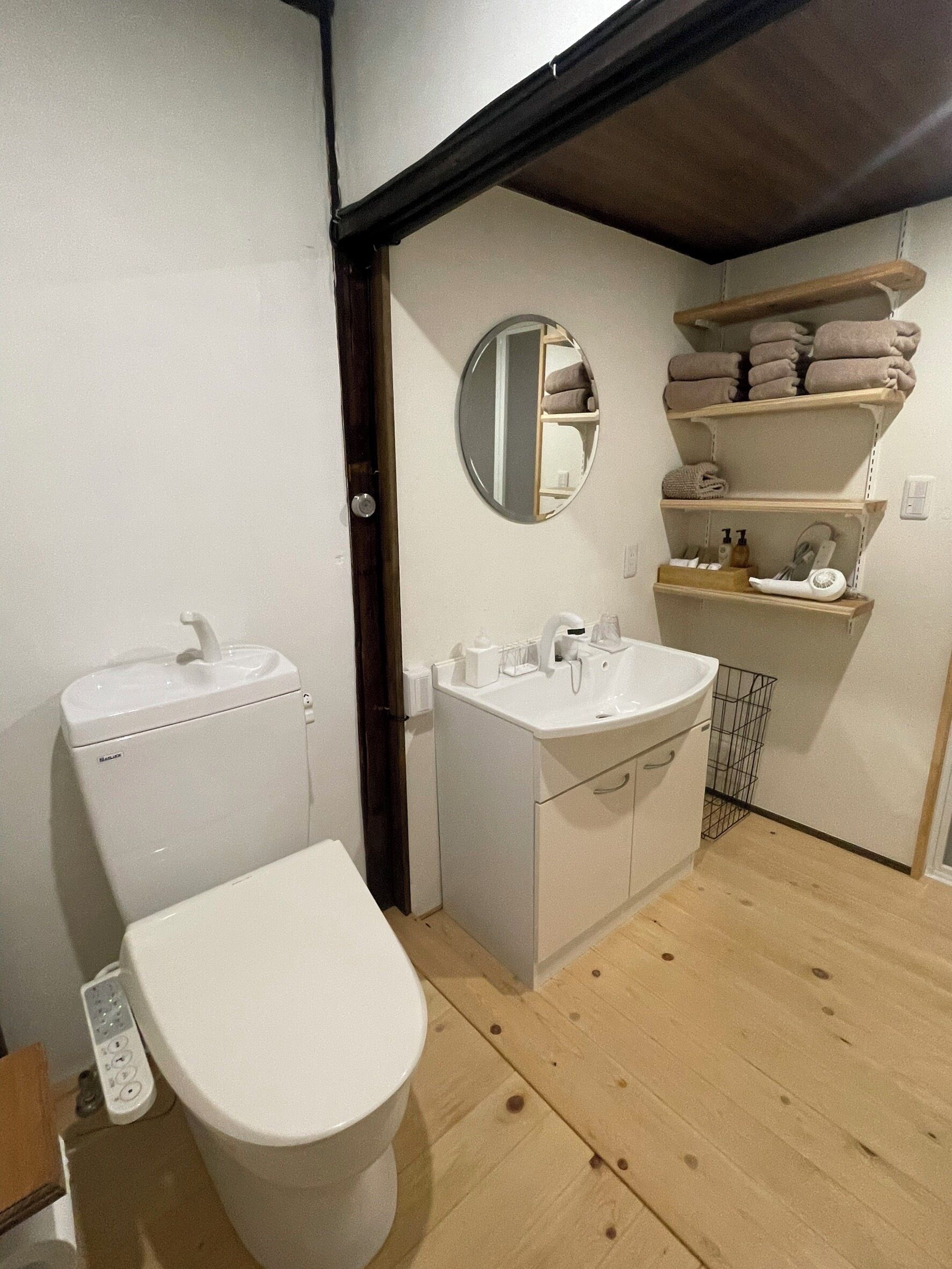 A small, compact bathroom with a toilet, sink, and mirror. There is a shelf with towels and toiletries.