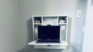 A room with a white desk, a laptop on a shelf, and a wooden floor.