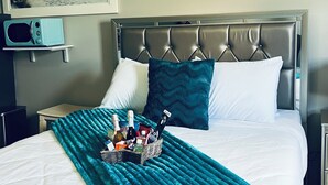 A neatly made bed with a teal throw blanket, a bedside table with a bottle of wine, and a framed landscape painting above the bed.