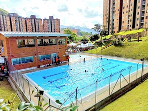 Una piscina rodeada por un edificio de ladrillo con varias ventanas, un área verde y un parque infantil.