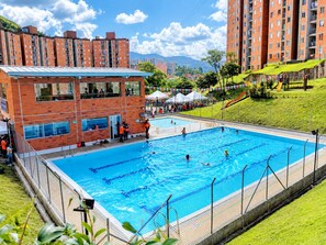 A swimming pool surrounded by a brick building with multiple windows, a grassy area with a slide, and a mountainous backdrop.