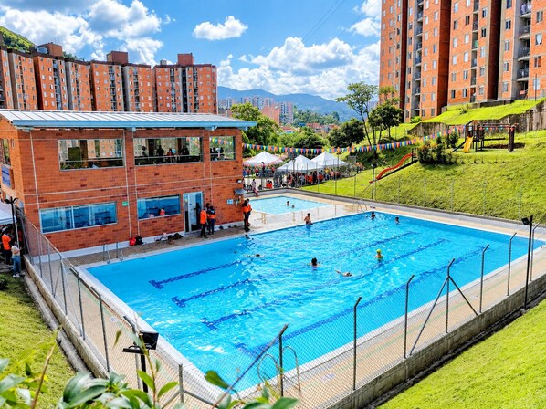 Una piscina rodeada por un edificio de ladrillo con múltiples ventanas, un área verde con un tobogán y montañas al fondo.