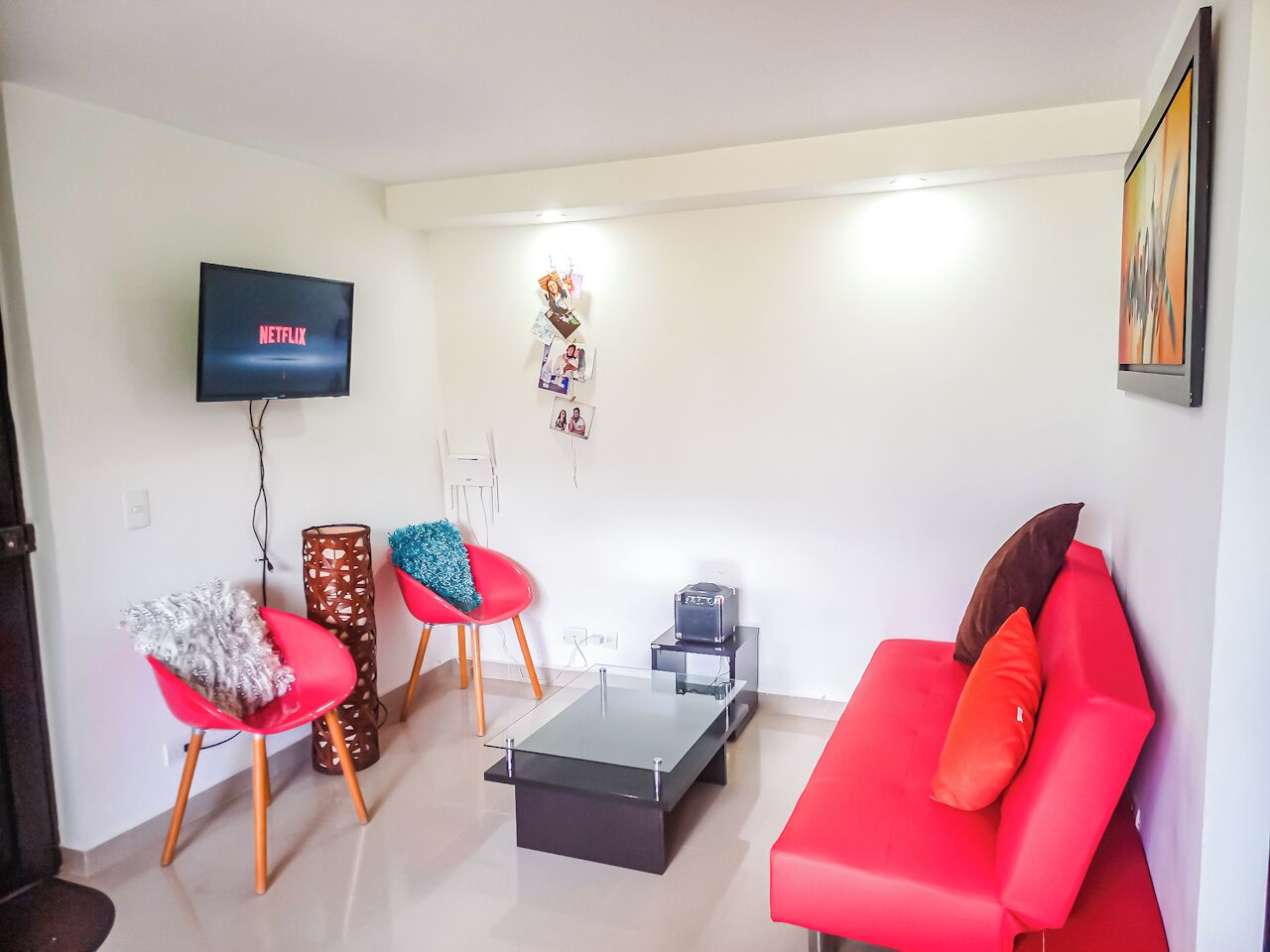 Una sala de estar con un sofá rojo, dos sillones rojos, una mesa de centro de vidrio, un televisor mostrando 'NETFLIX' y una estantería con fotografías.