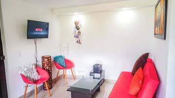 Una sala de estar con un sofá rojo, dos sillones rojos, una mesa de centro de vidrio, un televisor mostrando 'NETFLIX' y una estantería con fotografías.