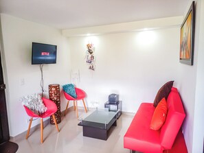 Una sala de estar con un sofá rojo, dos sillones rojos, una mesa de centro de vidrio, un televisor mostrando 'NETFLIX' y una estantería con fotografías.