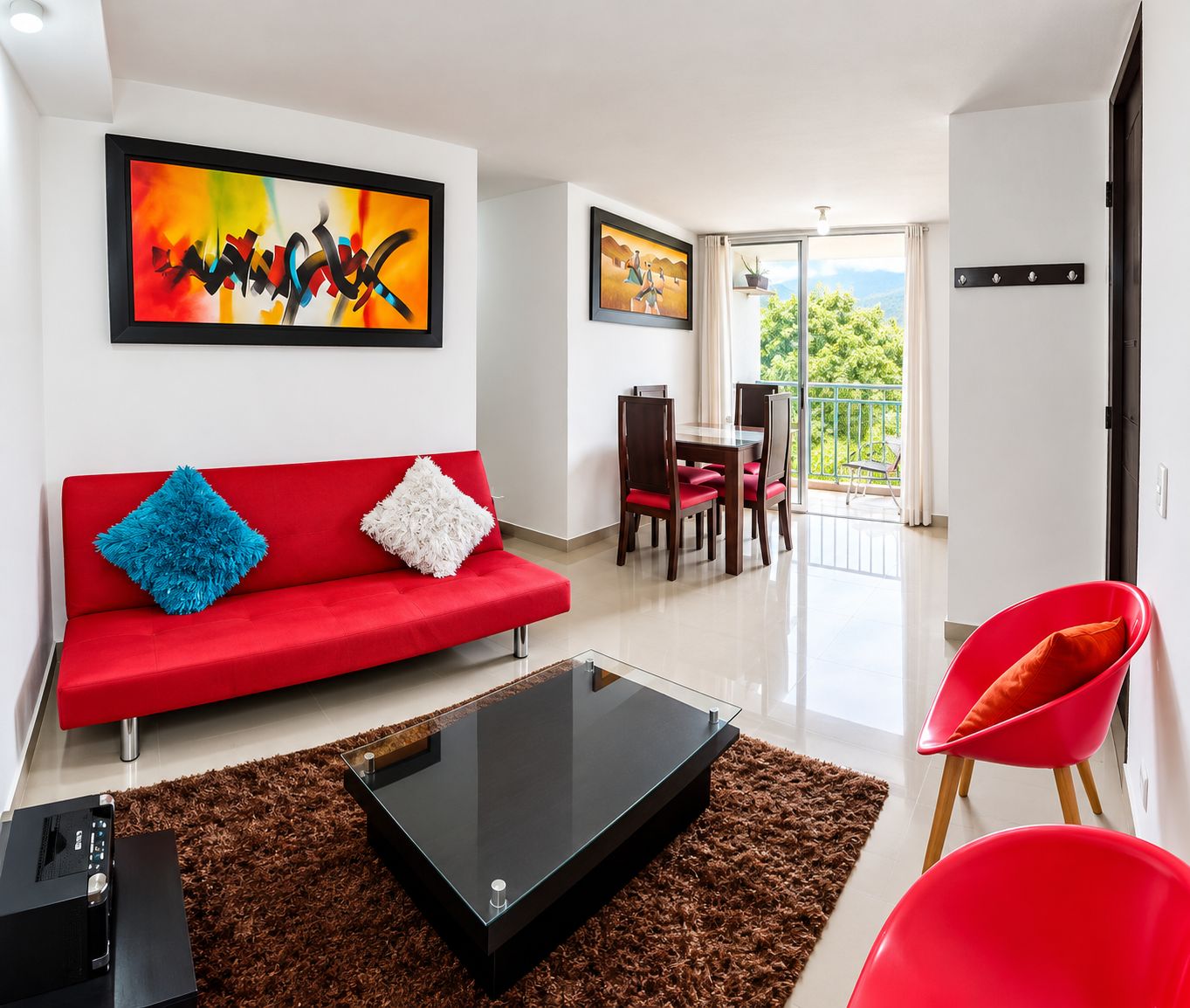 A modern living room with a red sofa, a glass coffee table, and a red chair. There are two framed artworks on the wall and a balcony with a view.