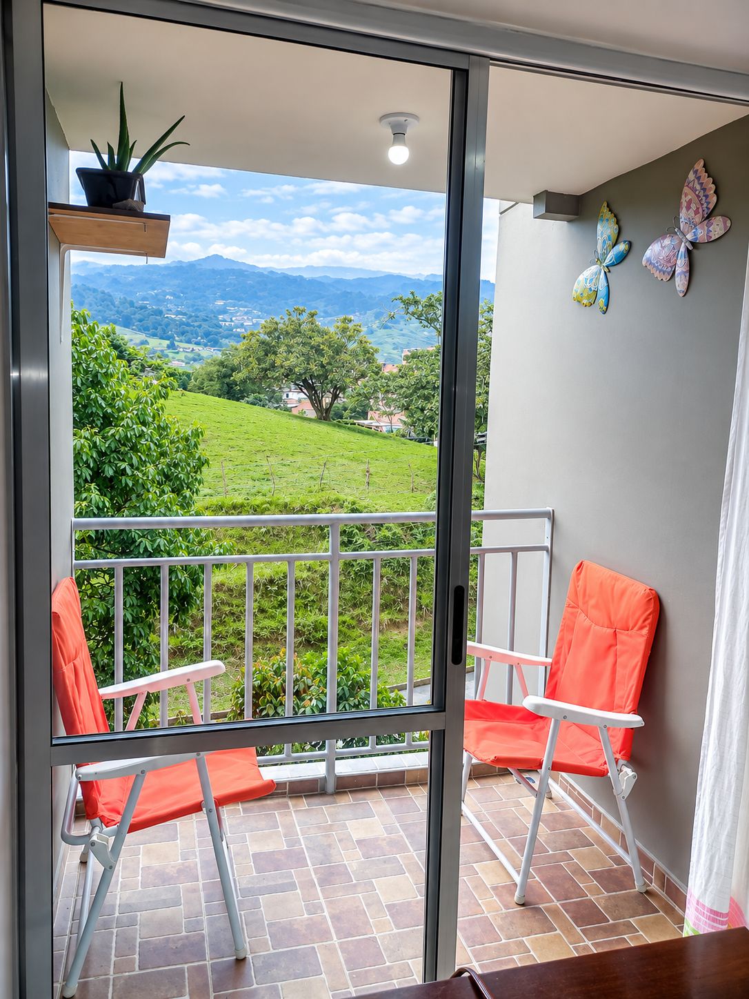 Varanda com vista para paisagem verde e montanhas, duas cadeiras, planta em vaso e arte decorativa de borboletas na parede.