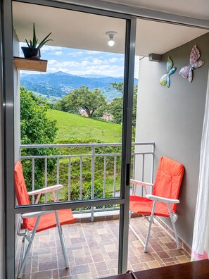 Un balcon donnant sur un paysage verdoyant et des montagnes, avec deux chaises, une plante en pot et une décoration murale représentant des papillons.