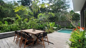 Eine Holzterrasse mit Tisch und Stühlen, umgeben von üppiger Vegetation und einem Schwimmbecken.