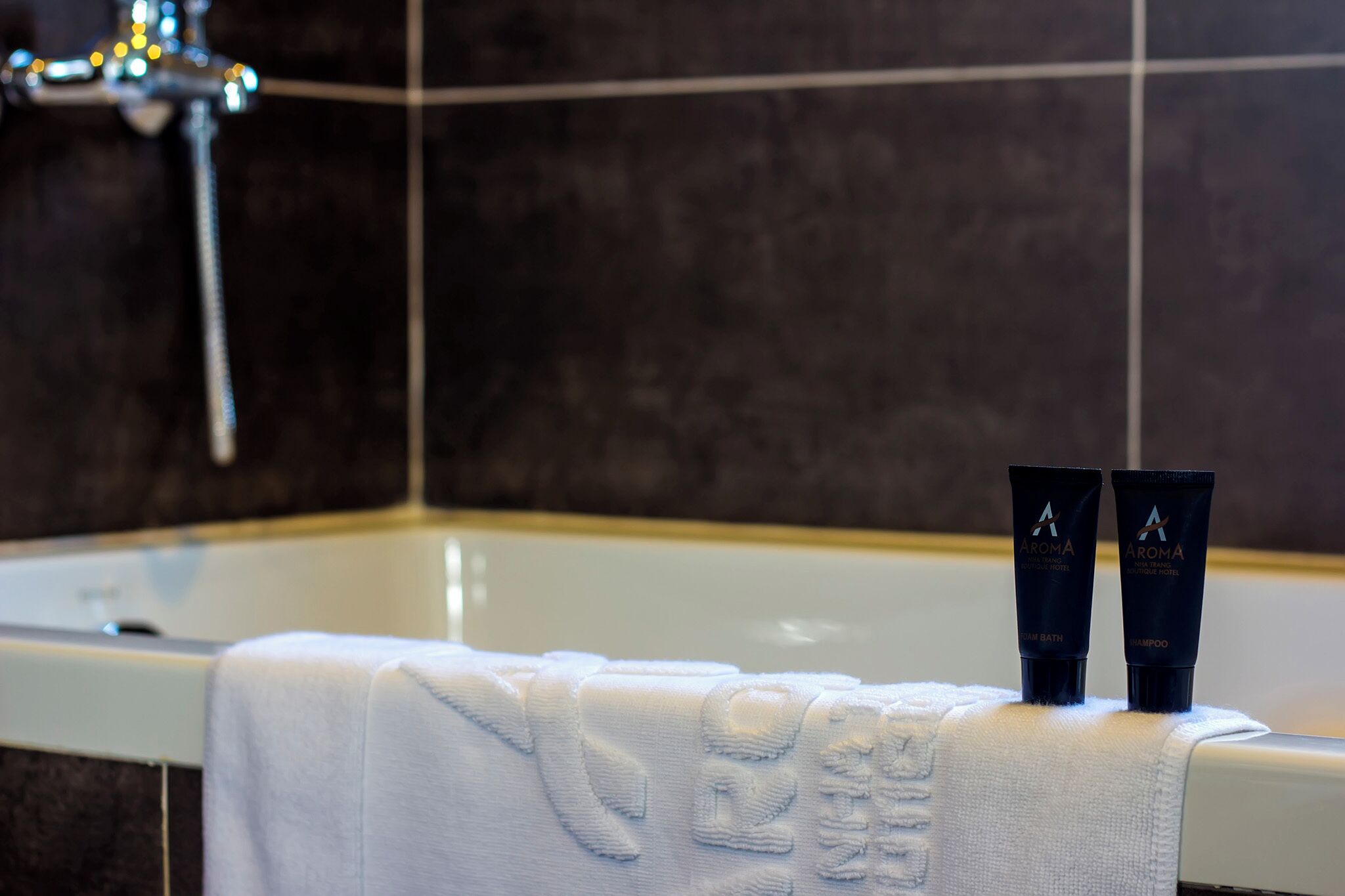 Une salle de bain avec une baignoire, deux bouteilles de produits de bain parfumés de la marque Aroma, et une serviette posée sur le bord.