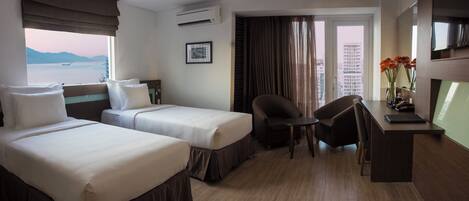 A hotel room with two beds, a desk, a chair, a TV, and a view of the sea.