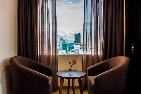 A room with two brown armchairs, a small round table with a vase of flowers, and a view of a cityscape through large windows with curtains. - Ocean View Nha Trang (Nha Trang)
