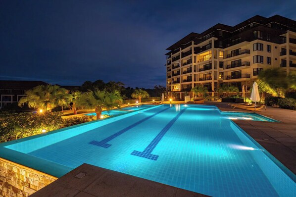 A swimming pool with a tiled deck, surrounded by palm trees and a multi-story building with balconies.