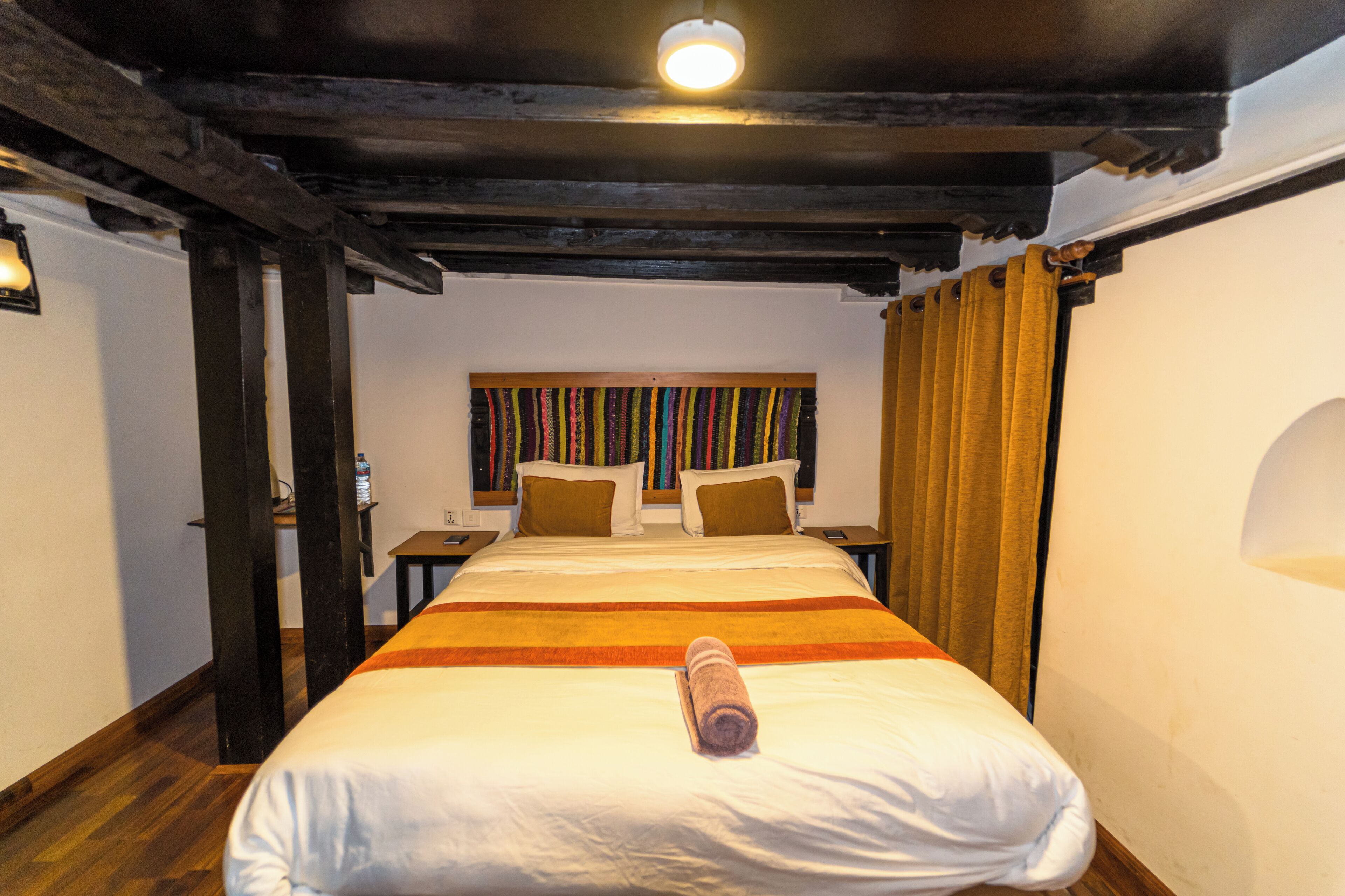 Habitación de hotel con cama, vigas de madera, una estantería y una ventana con cortinas.