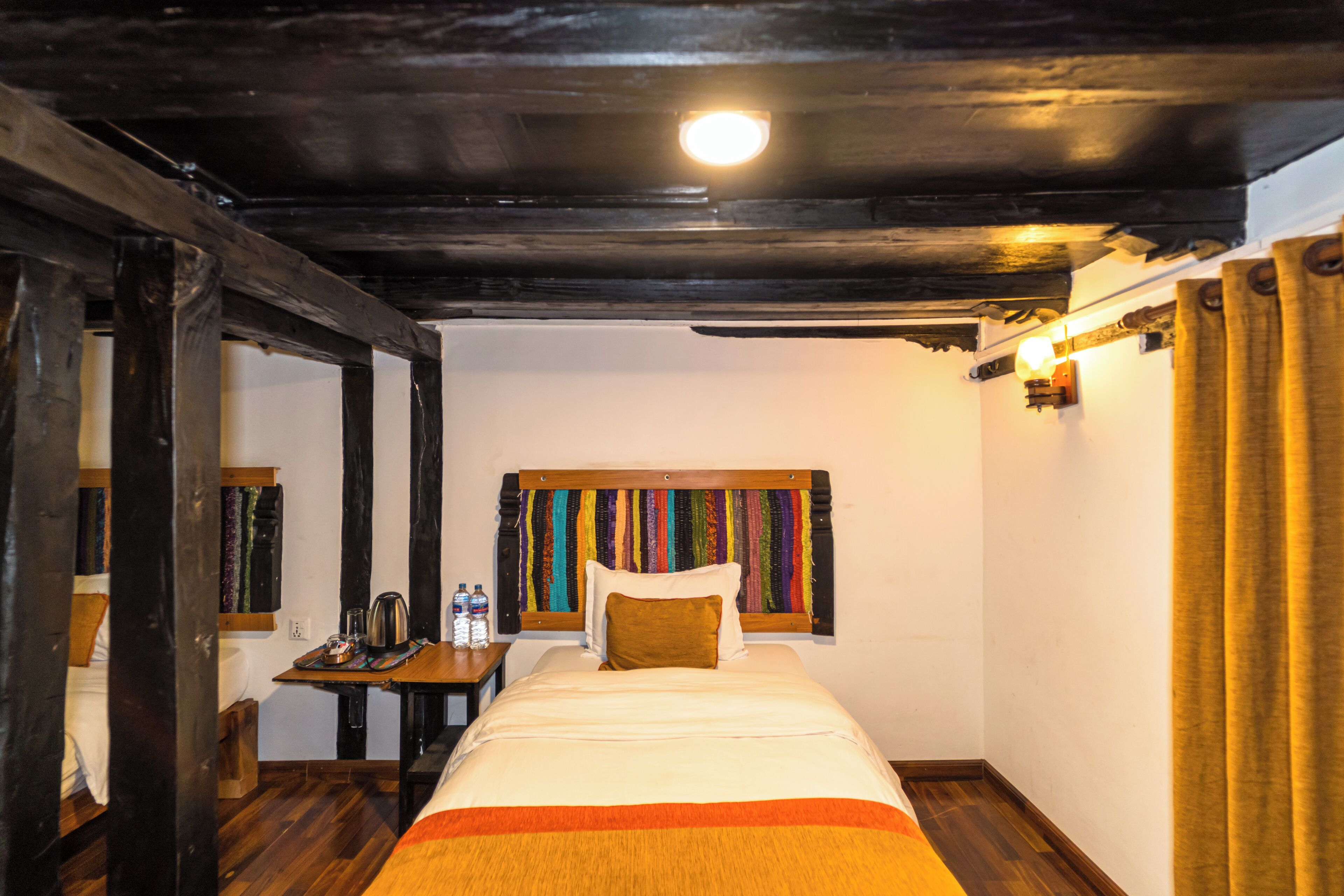 Una habitación con cama, vigas de madera, una mesa con hervidor, botellas y un tapiz de colores en la pared.