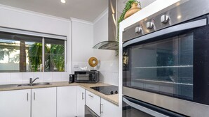 A modern kitchen with white cabinets, a stainless steel oven, a microwave, and a window with a view of palm trees. - Driftwood Mantaray Two Bedroom Two Bathroom Penthouse (Port Douglas)