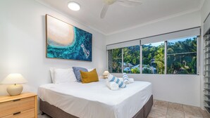 A bedroom with a bed, a nightstand, a painting, and a window with a view of greenery. - Driftwood Mantaray Two Bedroom Two Bathroom Penthouse (Port Douglas)