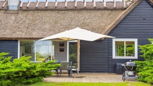 Une maison avec un toit de chaume, une terrasse avec une table et des chaises, et un barbecue.
