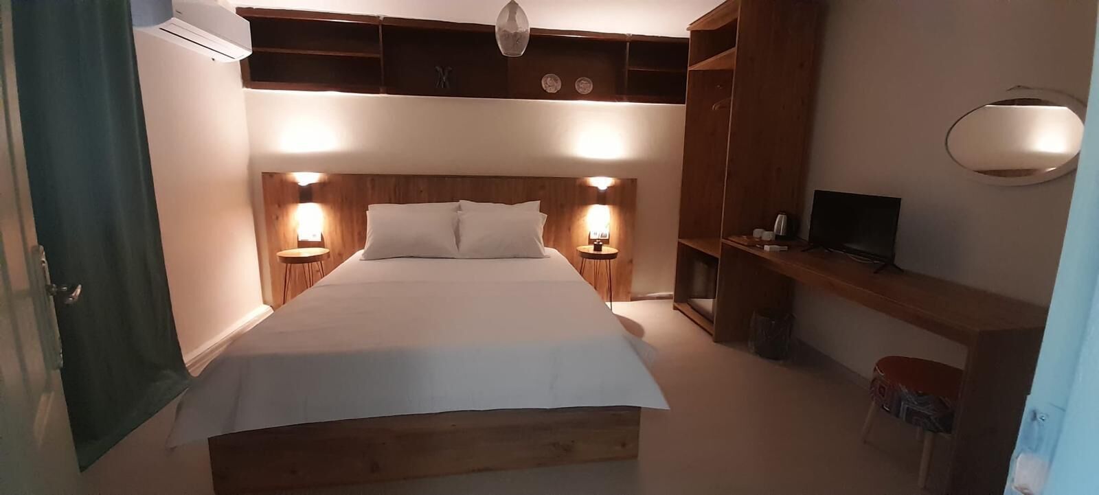 Un dormitorio con un cabecero de madera, dos mesitas de noche con lámparas, un televisor sobre una estantería de madera y una puerta a la izquierda.