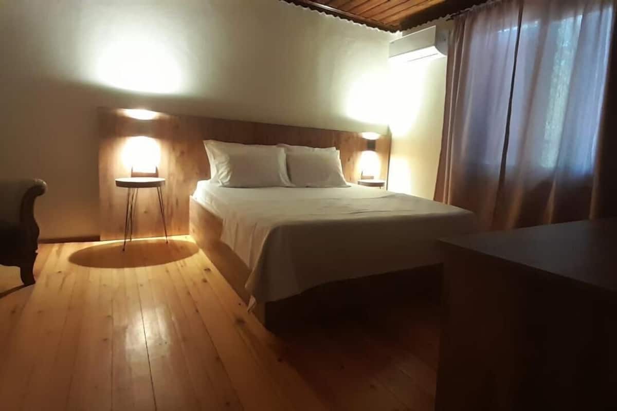 A room with a wooden floor, a bed with white bedding, two bedside lamps, a chair, and a coffee maker on a table.