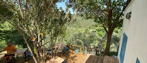 An outdoor wooden deck with tables and chairs, surrounded by trees and a view of a hilly landscape.