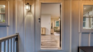 Una terraza de madera con un banco, una puerta que da a un salón con un sofá y una mesa de centro, y una lámpara sobre la puerta.