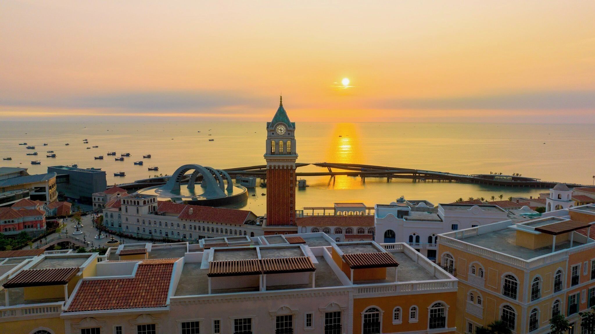 A coastal cityscape with a prominent clock tower, a bridge, and a calm sea at sunset.