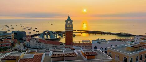 A coastal cityscape with a prominent clock tower, a bridge, and a calm sea at sunset.